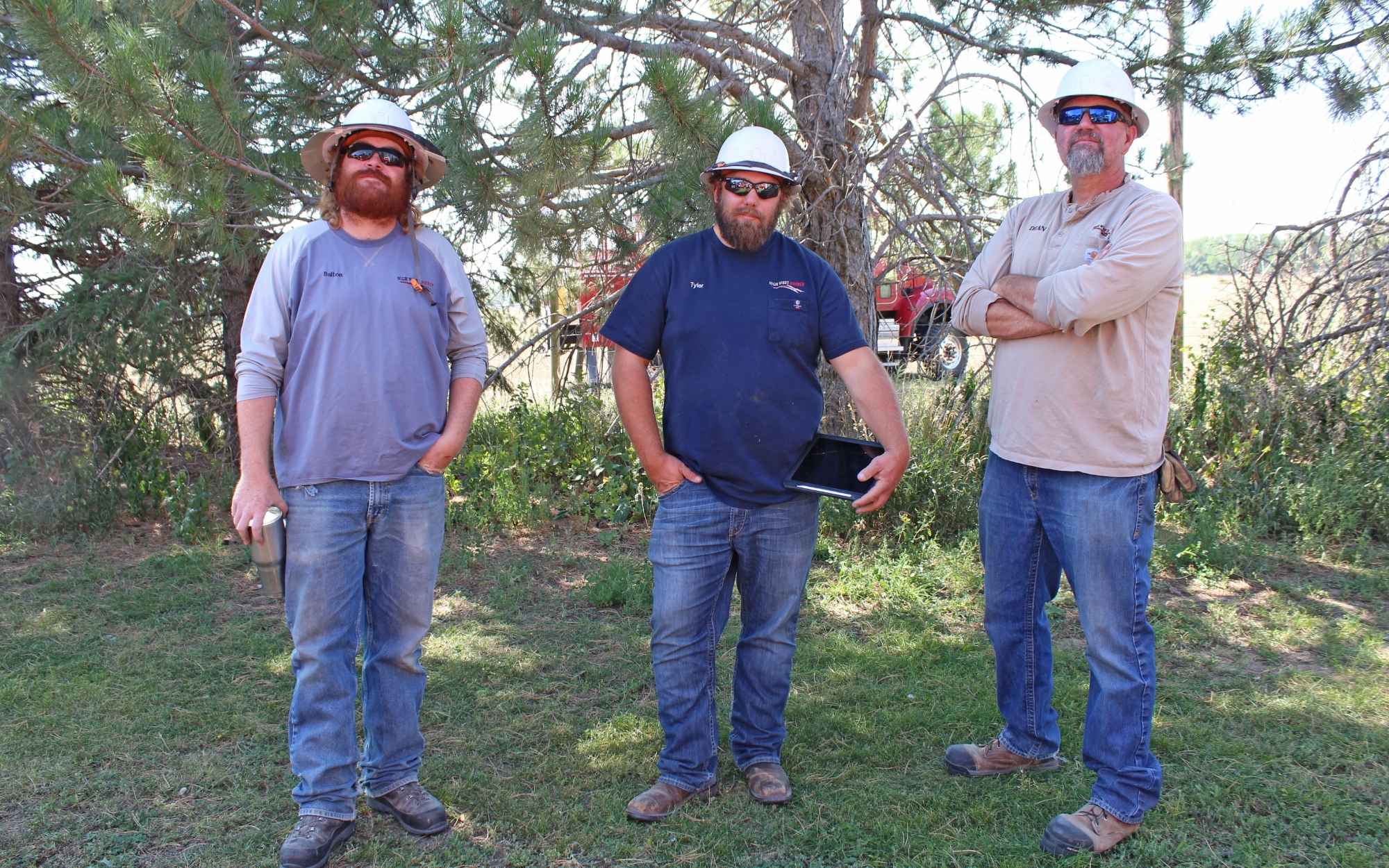 Three individuals wearing hard hats and work clothes stand outdoors in front of trees and brush. One holds a tablet.