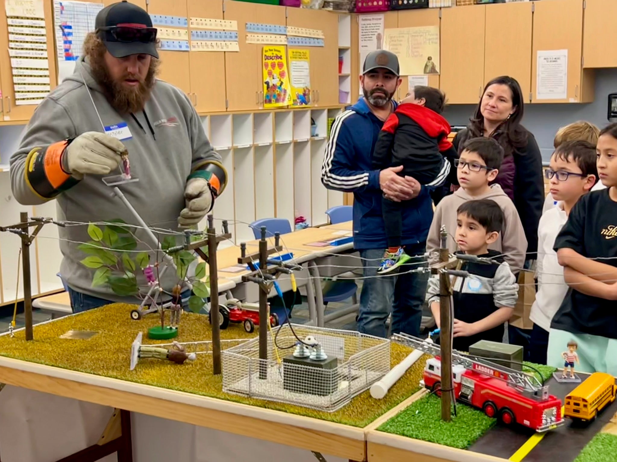 A presenter wearing protective gloves demonstrates a miniature electrical grid model to a group of children and adults in a classroom setting.
