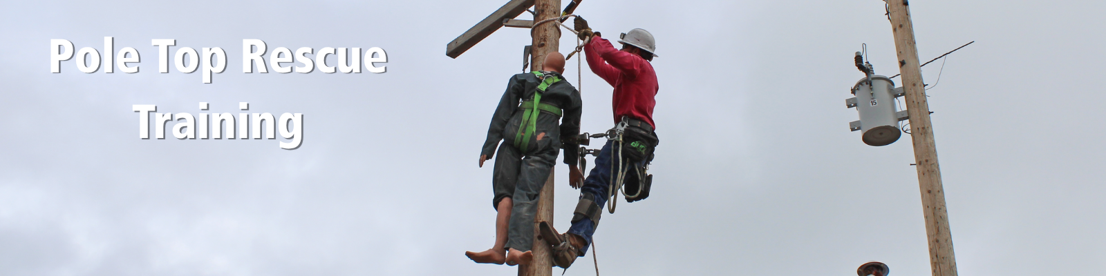 A utility worker in safety gear completing pole rescue training. The worker, wearing a red shirt and helmet, is above the dummy, who is wearing a dark jumpsuit with a green harness. Pole Top Rescue Training text on left side of image. 