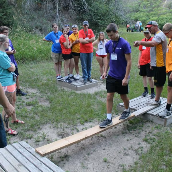 A group of participants stands outdoors in a grassy area watching a team-building activity. One participant carefully walks across a narrow wooden plank suspended between two small platforms while others observe from the sides. Several participants wear colorful T-shirts and name tags. Trees and additional people can be seen in the background, creating the setting of an outdoor leadership or youth camp event.