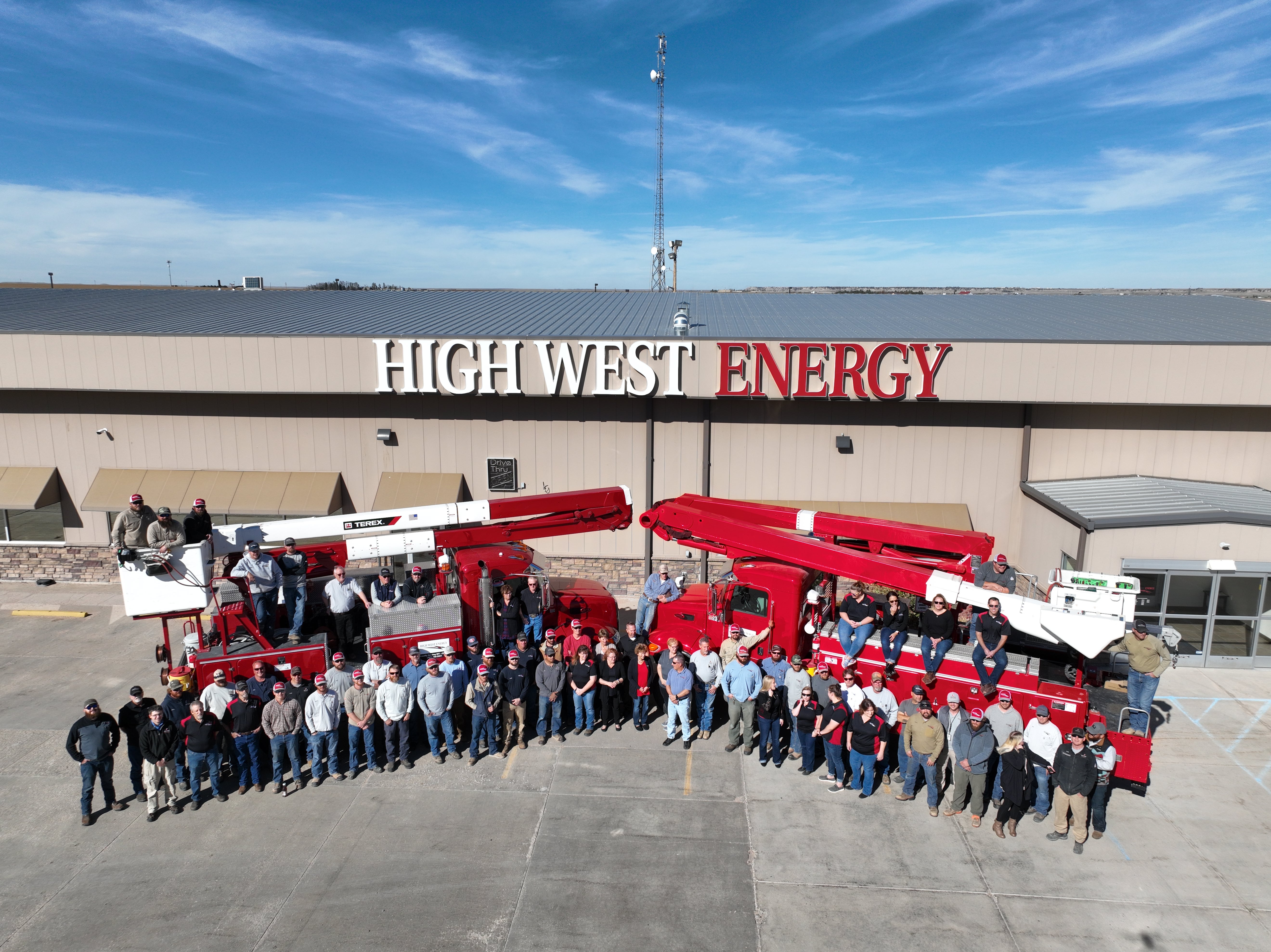 Aerial group photo of High West Energy employees gathered in front of the cooperative headquarters building, with two red bucket trucks parked in the foreground and the High West Energy sign displayed on the building.