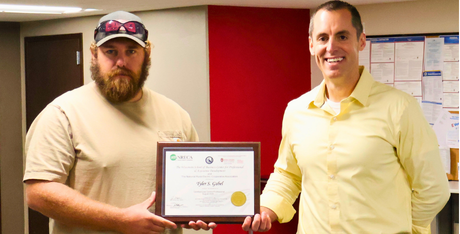 Two individuals standing indoors and holding a framed certificate together. The certificate is centered in the image and features a seal and official formatting. One individual is wearing a light-colored shirt and cap, while the other is wearing a yellow button-down shirt. The background includes a red accent wall, office bulletin boards, and a doorway. The image appears to document the presentation or recognition of a professional certification or achievement.
