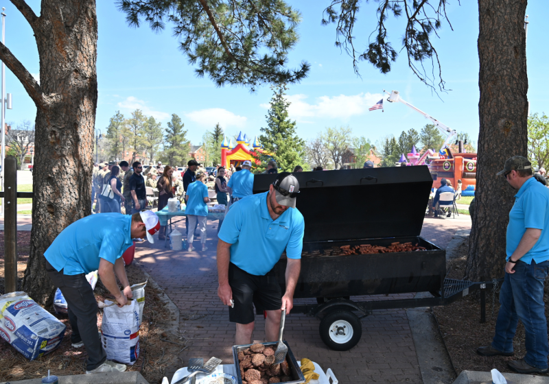 People in blue shirts gather around a large outdoor grill, preparing food with bags and trays of ingredients on the ground, while a crowd watches and flags fly in the background.