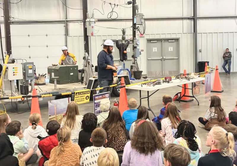 In a large workshop, a speaker stands beside a table of electronics while a group of children gathers behind yellow tape to watch a guided demonstration near machinery and cables.