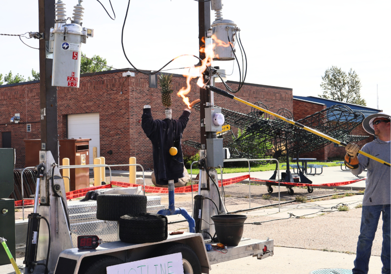 Electrical safety demonstration showing a utility safety trailer with transformers, power lines, and a mannequin used to demonstrate electrical hazards, including an energized power line contact simulation.