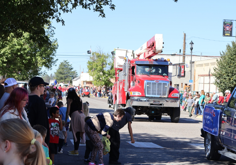A red-and-white truck leads a parade down a sunny street as people line the sidewalk.