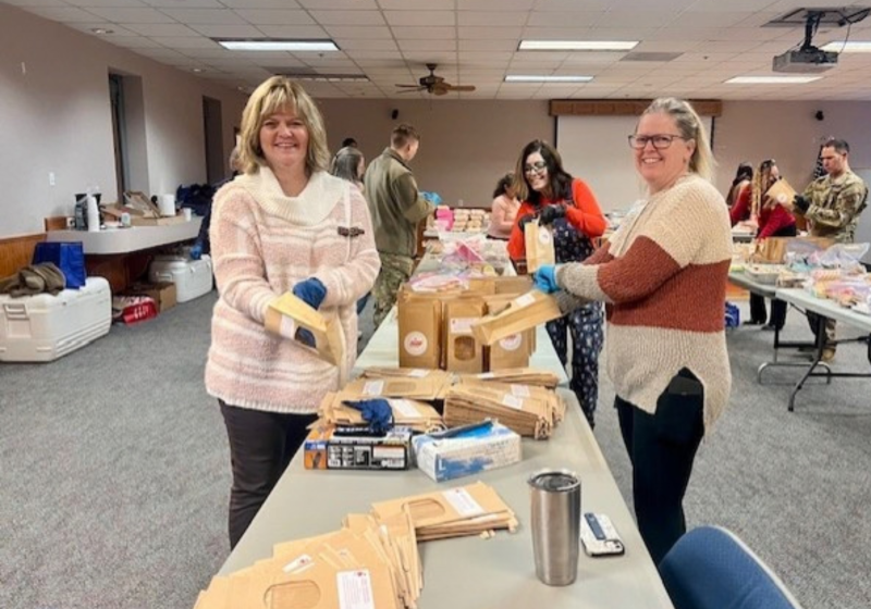 In a community room, two people stand beside a long table covered with packaged supplies and boxes, while others chat in the background during a volunteer or packing event.