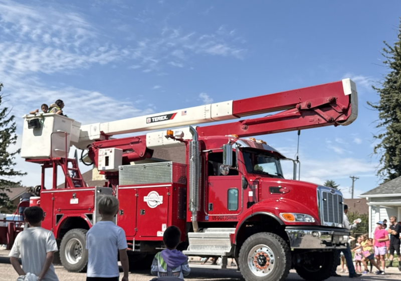A red and white utility truck with an extendable bucket is driving in a parade, with several people standing nearby and children watching.
