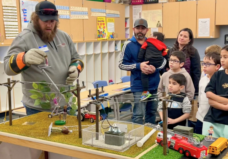 In a classroom makerspace, a bearded instructor in safety gear demonstrates a model science setup on a grassy table while several children in a line watch closely.