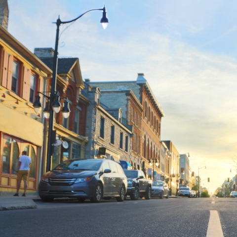 Sunset view down a quiet downtown street with parked cars on the side and brick storefronts lining the road under a pale blue sky.