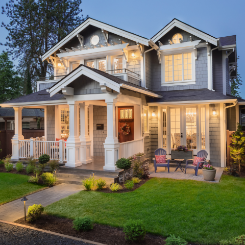 Two-story house with a front porch and lit windows at dusk, white columns and railings, landscaped lawn with shrubs and walkway, and outdoor seating on the right under a clear blue sky.